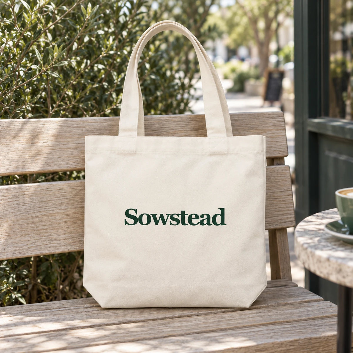 Natural Sowstead cotton tote on a timber bench, showing its forest-green front wordmark.